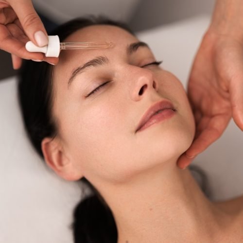 From above of crop unrecognizable beautician applying essential oil with pipette on face of relaxed young female client with dark hair, lying on table with closed eyes in beauty salon